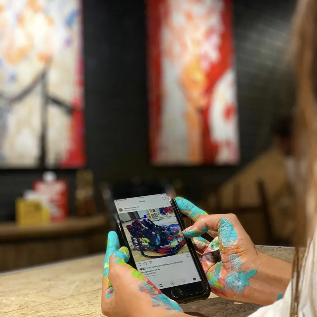 Tamy con las manos manchadas de pintura mirando el teléfono, con sus obras al fondo